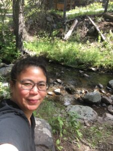 Portrait of the artist outdoors beside a shallow creek in a wooded area, photographed on location for ACFA Creative House.