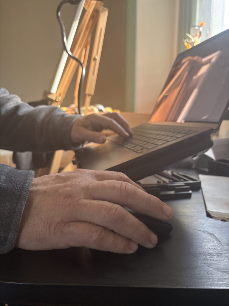 Engineer workspace at ACFA Creative House showing hands working on a laptop at a raised desk with task lighting and tools nearby, representing systems design and technical writing.